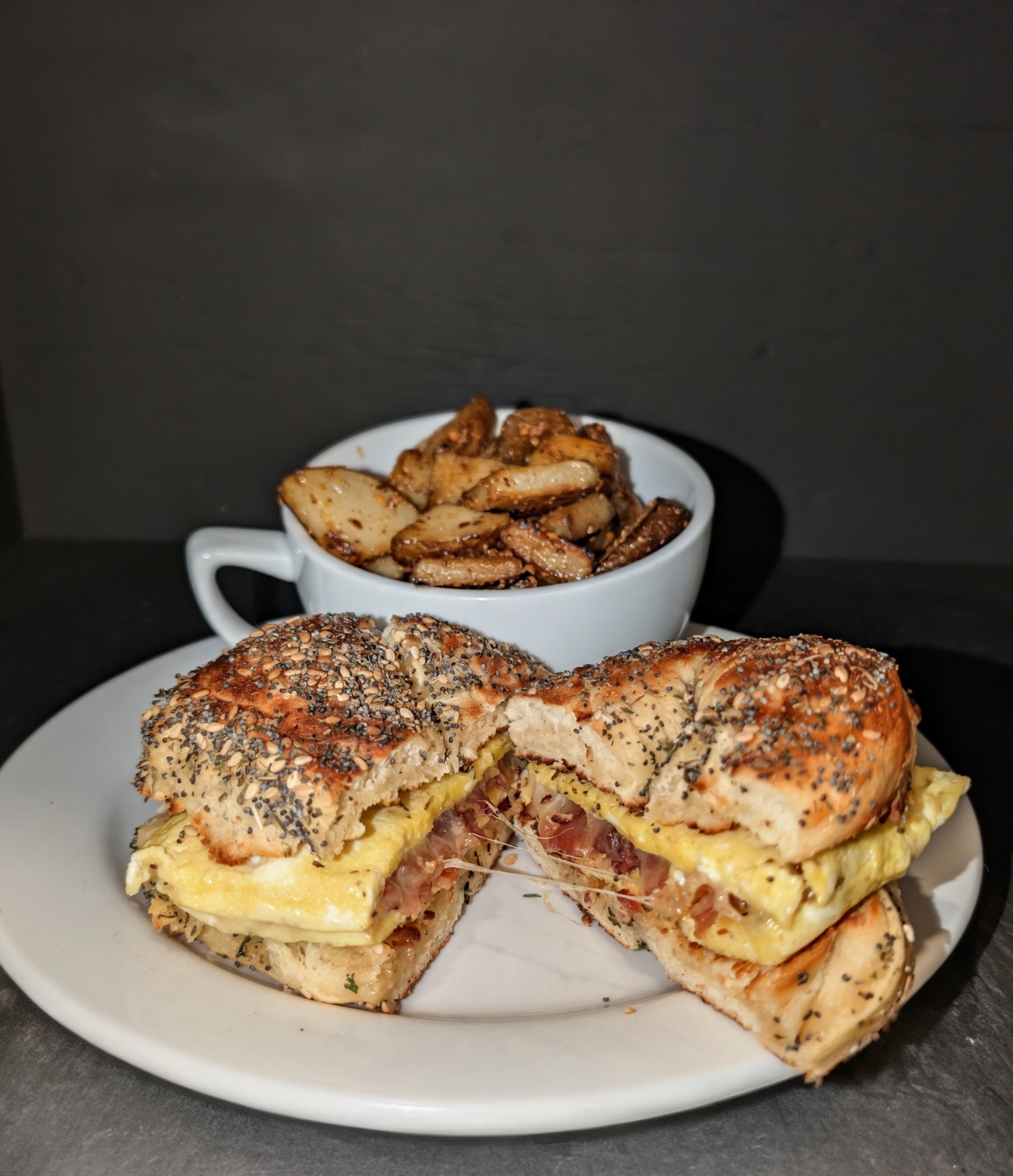 An egg, bacon and cheese sandwich on an everything bagel, with a cup of seasoned potatoes behind it