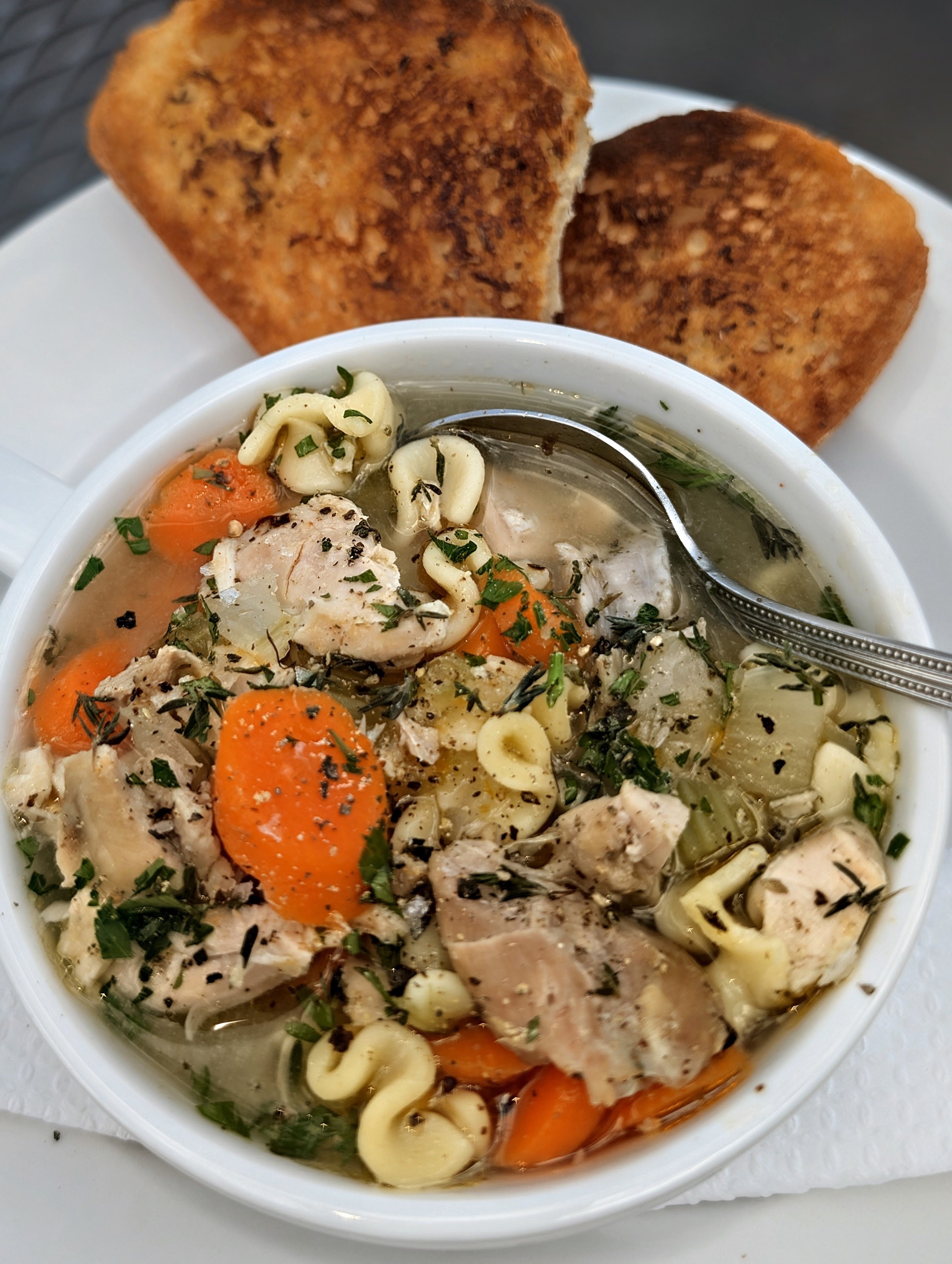 A bowl of chicken noodle soup with carrots and herbs, a spoon resting in it and griddled toast behind