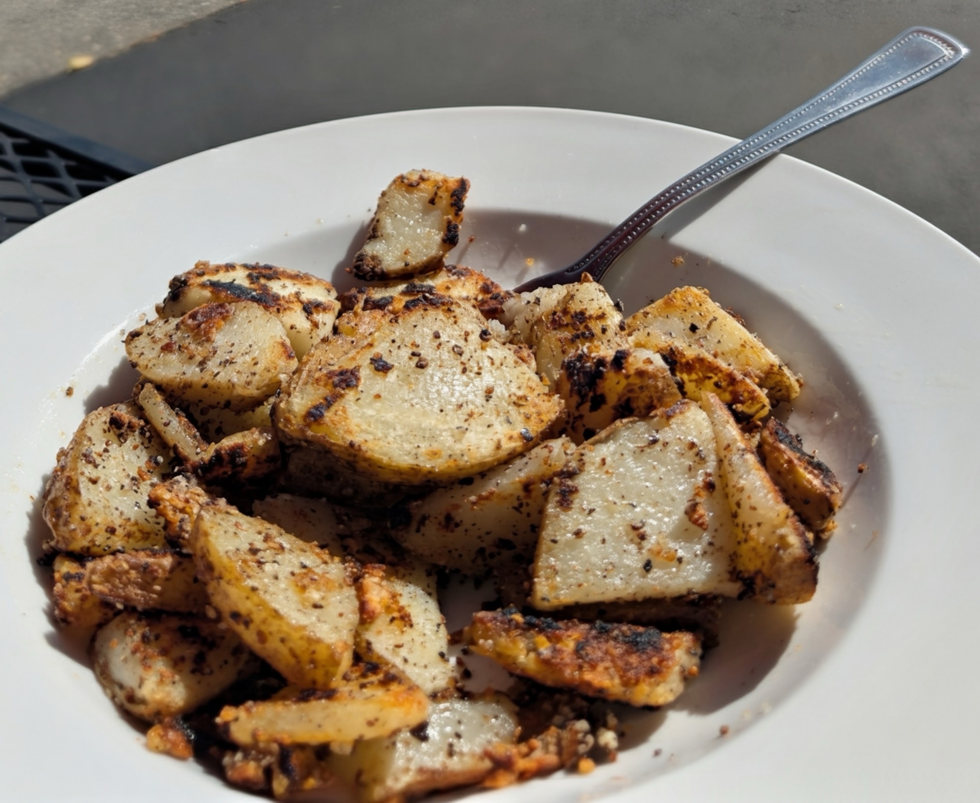A plate of golden, seasoned potato chunks with a fork, on a table in the sun