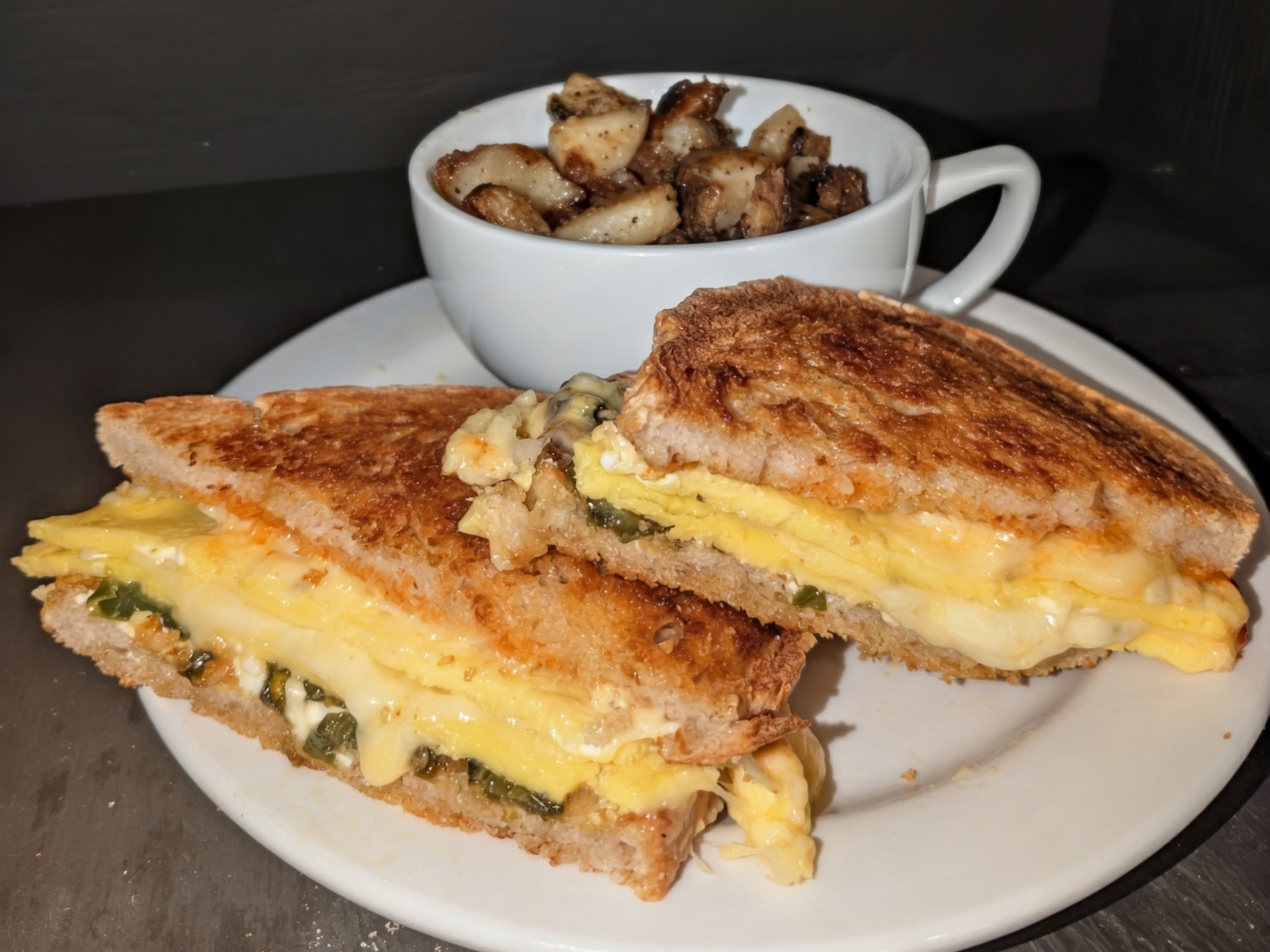 A griddled egg and cheese sandwich with green jalapeños, cut in half, with a cup of potatoes